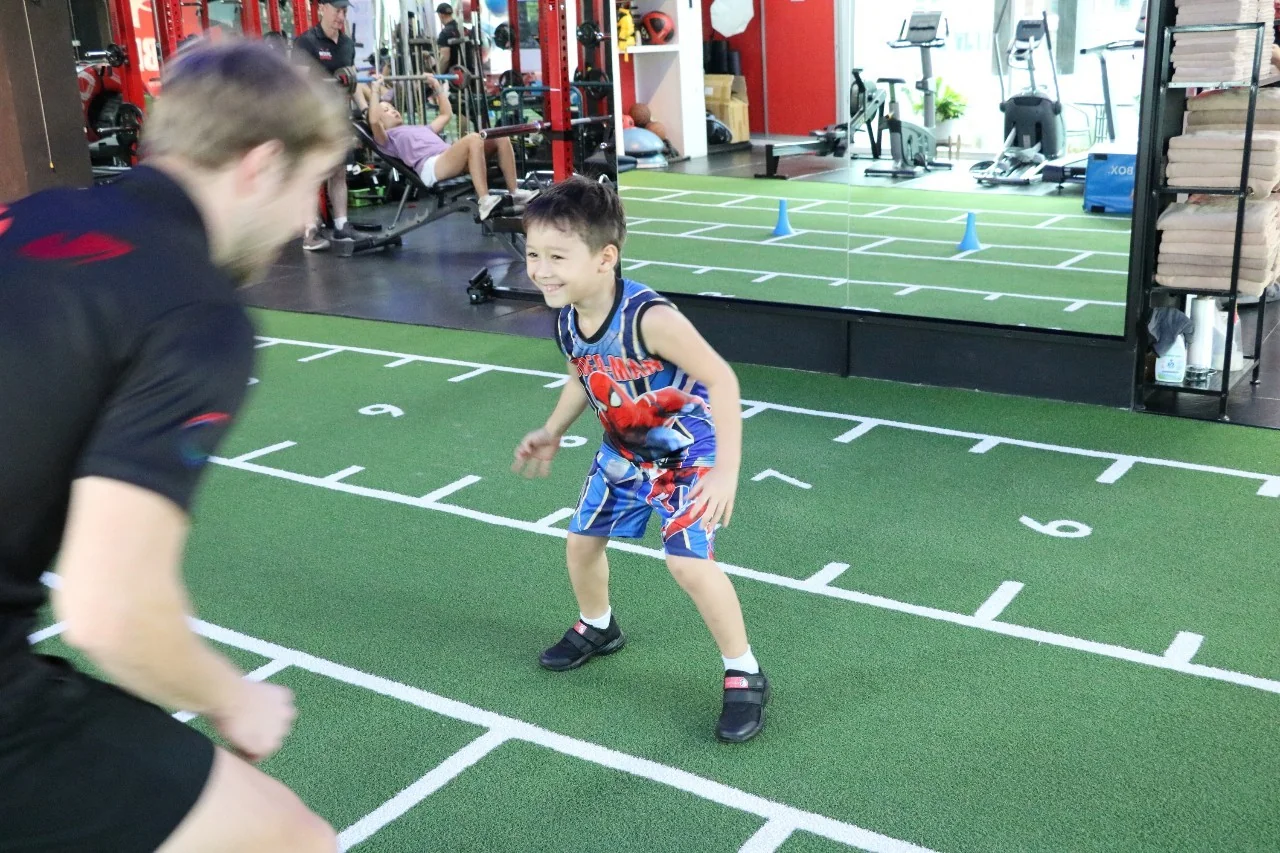 Young boy smiling during a one-to-one agility drill with a Body By Beast coach on the studio turf in Bangkok